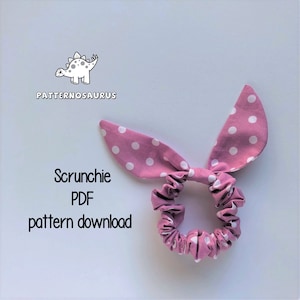 May include: A pink scrunchie with white polka dots and bunny ear bows. The scrunchie is made of fabric and has a gathered look. The text "Scrunchie PDF pattern download" is printed below the scrunchie.