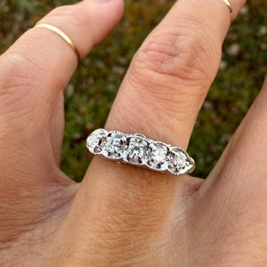 May include: A silver ring with five clear stones set in a row. The ring is on a person's finger, with a gold band on the adjacent finger. The background is blurred green.