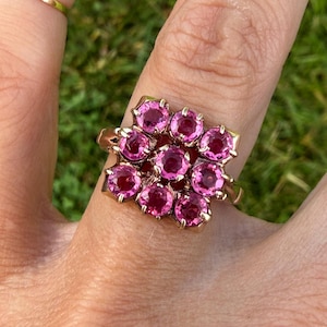 May include: A gold ring featuring a cluster of nine round, faceted, vibrant pink gemstones arranged in a square formation. The stones are held in place by small metal prongs, creating a dense, floral-like pattern on the top of the ring band.
