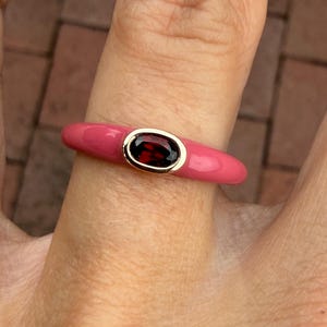 May include: A ring featuring a smooth, glossy pink enamel band with a central, horizontally oriented oval-shaped dark red gemstone set in a polished gold-toned metal bezel. The design is simple and uniform, showcasing a vibrant contrast between the pink enamel and the deep red stone.