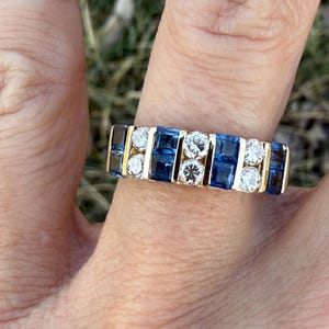 May include: A gold ring with alternating rows of blue and clear gemstones. The ring has a rectangular design with the gemstones set in a channel setting. The ring is worn on a finger, with a gold band visible on the adjacent finger.