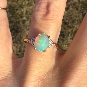 Puede incluir: Un anillo de oro con un &oacute;palo ovalado en el centro. La piedra muestra destellos iridiscentes de luz verde y naranja. El anillo presenta un peque&ntilde;o grupo de tres piedras transparentes de corte redondo a cada lado del engaste central.