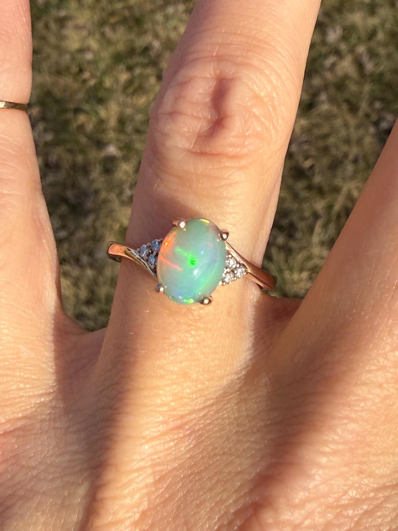 Puede incluir: Un anillo de oro con un &oacute;palo ovalado en el centro. La piedra muestra destellos iridiscentes de luz verde y naranja. El anillo presenta un peque&ntilde;o grupo de tres piedras transparentes de corte redondo a cada lado del engaste central.