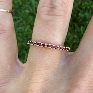May include: A thin gold band ring featuring a continuous row of small, round, deep red garnet gemstones set in a prong setting. The ring is worn on a finger, showcasing the uniform size and circular shape of the stones along the entire band.