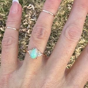 Puede incluir: Un anillo de oro con una piedra central de &oacute;palo ovalada e iridiscente, flanqueada por tres peque&ntilde;as piedras transparentes a cada lado. Se llevan dos anillos de oro finos y lisos adicionales en los dedos adyacentes.
