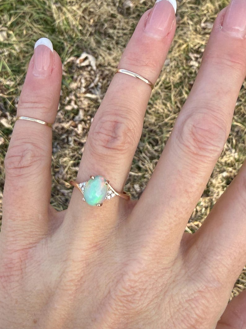 Puede incluir: Un anillo de oro con una piedra central de &oacute;palo ovalada e iridiscente, flanqueada por tres peque&ntilde;as piedras transparentes a cada lado. Se llevan dos anillos de oro finos y lisos adicionales en los dedos adyacentes.