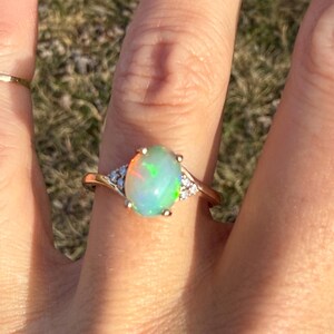 Puede incluir: Un anillo de oro con una piedra preciosa de &oacute;palo ovalada en el centro que muestra destellos iridiscentes verdes y naranjas. La banda est&aacute; decorada con peque&ntilde;as piedras transparentes a ambos lados, creando un aspecto delicado y refinado.