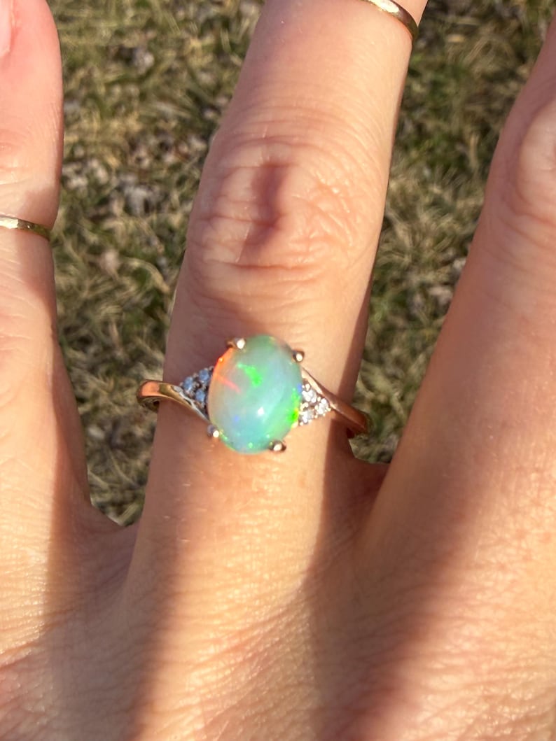 Puede incluir: Un anillo de oro con una piedra preciosa de &oacute;palo ovalada en el centro que muestra destellos iridiscentes verdes y naranjas. La banda est&aacute; decorada con peque&ntilde;as piedras transparentes a ambos lados, creando un aspecto delicado y refinado.