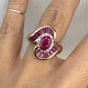 Peut inclure: Une bague en or avec une pierre centrale en rubis ovale, entourée de diamants et de rubis taille baguette. La bague présente un design tourbillonnant unique et un anneau en or poli.