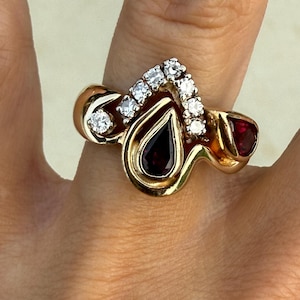 May include: A gold ring featuring a central pear-shaped dark red gemstone, a smaller triangular red gemstone on the side, and a chevron-shaped row of five small round clear diamonds set above the central stone, with a smooth polished gold band.