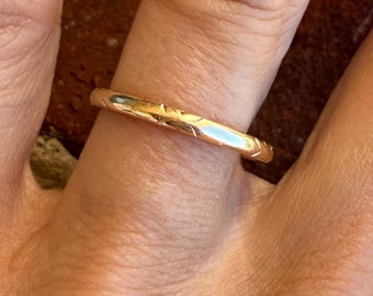 Puede incluir: Un anillo de banda fina de oro rosa pulido que presenta una serie de pequeños patrones grabados en forma de cheurón o V, espaciados uniformemente alrededor de la superficie exterior del metal, creando un detalle texturizado sutil.