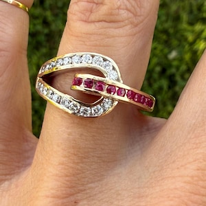Könnte beinhalten: Ein Goldring mit einem offenen, ineinandergreifenden Schleifendesign. Ein Teil des Rings ist mit einer Reihe klarer, rund geschliffener Steine besetzt, während ein kleinerer, überlappender horizontaler Balken mit tiefroten Steinen besetzt ist.
