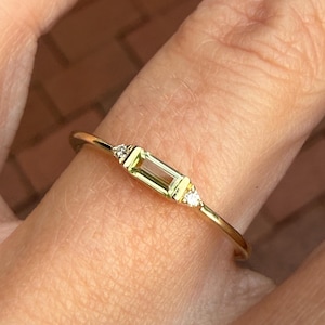 May include: A delicate gold ring featuring a rectangular, light green gemstone as the centerpiece. Two small, round, clear gemstones flank the main stone. The ring is set on a slender gold band, showcasing a minimalist design.