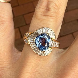 Puede incluir: Un anillo de oro con una gran gema azul claro en forma de corazón en el centro, rodeada de gemas claras rectangulares más pequeñas. La banda del anillo también es de oro, con pequeñas gemas claras adicionales.