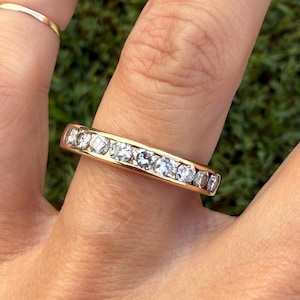 May include: A gold band ring featuring a channel setting with a row of nine round, clear, faceted stones. The ring has a polished finish and is worn on a finger, with a thin, plain gold band visible on an adjacent finger.