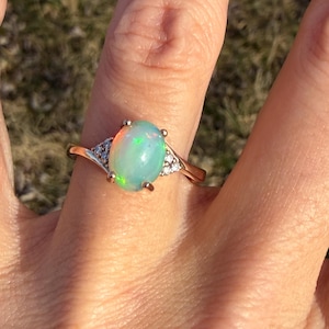 Puede incluir: Un anillo con un &oacute;palo cabuj&oacute;n de forma ovalada montado en una banda de oro pulido. El &oacute;palo central muestra destellos iridiscentes verdes y naranjas. Tres peque&ntilde;as piedras transparentes de talla redonda est&aacute;n engastadas a cada lado.