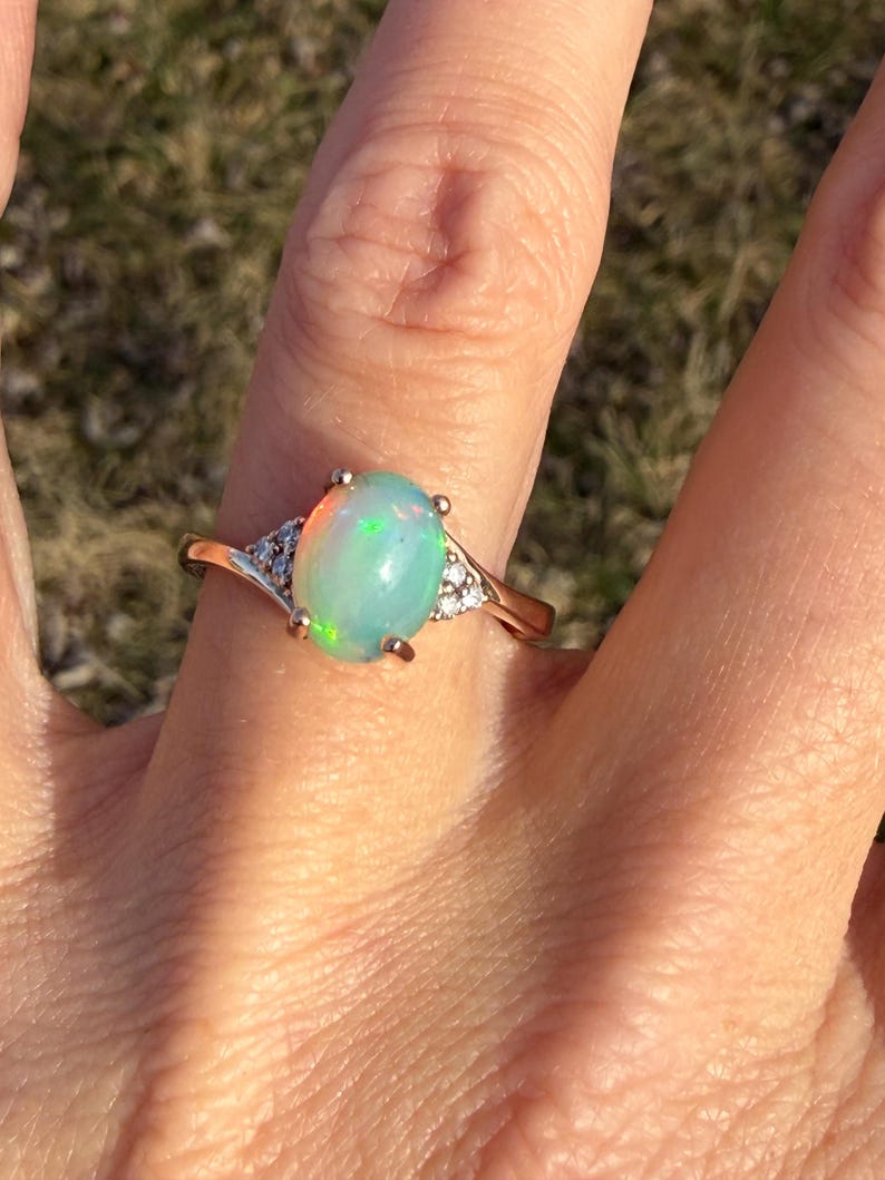 Puede incluir: Un anillo con un &oacute;palo cabuj&oacute;n de forma ovalada montado en una banda de oro pulido. El &oacute;palo central muestra destellos iridiscentes verdes y naranjas. Tres peque&ntilde;as piedras transparentes de talla redonda est&aacute;n engastadas a cada lado.