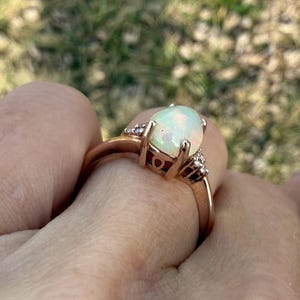 Puede incluir: Un anillo de oro rosa con un &oacute;palo central ovalado en cabuj&oacute;n con destellos iridiscentes. El engaste incluye peque&ntilde;as piedras transparentes a ambos lados del &oacute;palo, sujetas por una montura de cuatro garras sobre una banda lisa y pulida.