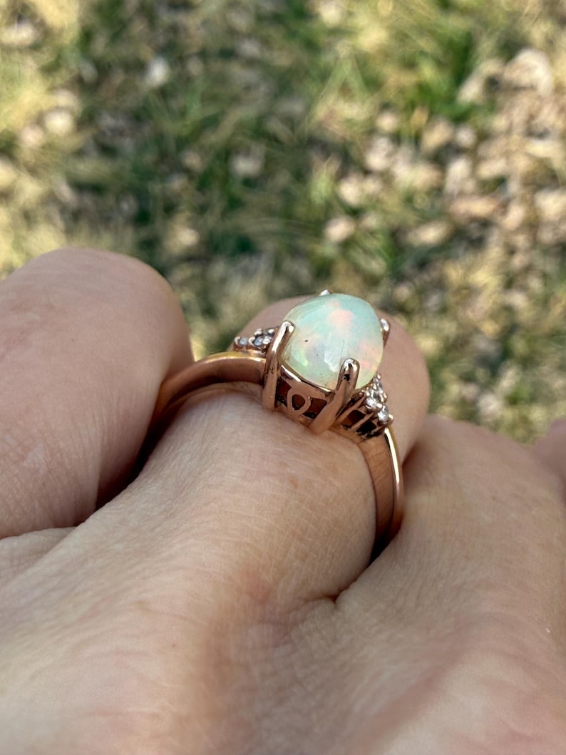 Puede incluir: Un anillo de oro rosa con un &oacute;palo central ovalado en cabuj&oacute;n con destellos iridiscentes. El engaste incluye peque&ntilde;as piedras transparentes a ambos lados del &oacute;palo, sujetas por una montura de cuatro garras sobre una banda lisa y pulida.