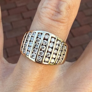 May include: A gold-toned ring with multiple rows of small, clear gemstones. The ring is wide and rectangular, with the gemstones set in parallel lines across the top. The ring is on a finger.