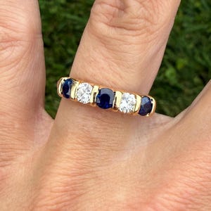 May include: A gold band ring with alternating dark blue sapphires and clear diamonds. The ring has five stones, set in a channel setting. The ring is displayed on a finger. The gemstones are round-cut.