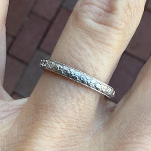 May include: A silver ring with a floral pattern. The ring is worn on a finger, showcasing the intricate details of the design. The ring's surface is adorned with a repeating pattern of small, stylized flowers, creating a delicate and elegant look.