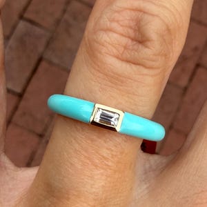May include: A bright turquoise enamel ring featuring a single, clear, baguette-cut stone set in a gold-toned metal bezel. The band has a smooth, rounded profile and a vibrant, solid color finish that contrasts with the central rectangular gemstone.