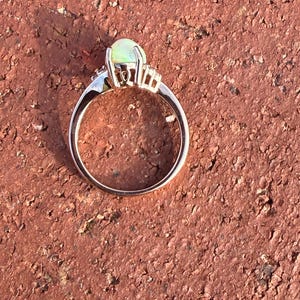Puede incluir: Un anillo con un &oacute;palo central de corte cabuj&oacute;n con un brillo p&aacute;lido e iridiscente, montado en una banda de metal pulido. El engaste incluye peque&ntilde;as piedras claras a los lados del &oacute;palo principal, aseguradas por varias garras.