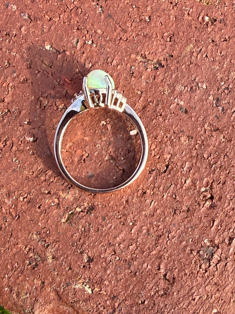 Puede incluir: Un anillo con un &oacute;palo central de corte cabuj&oacute;n con un brillo p&aacute;lido e iridiscente, montado en una banda de metal pulido. El engaste incluye peque&ntilde;as piedras claras a los lados del &oacute;palo principal, aseguradas por varias garras.