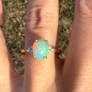 Puede incluir: Un anillo con un cabuj&oacute;n de &oacute;palo ovalado con destellos iridiscentes en tonos verde, naranja y azul. El &oacute;palo est&aacute; engastado en una banda de oro pulido, decorada con tres peque&ntilde;as piedras redondas y transparentes a cada lado.