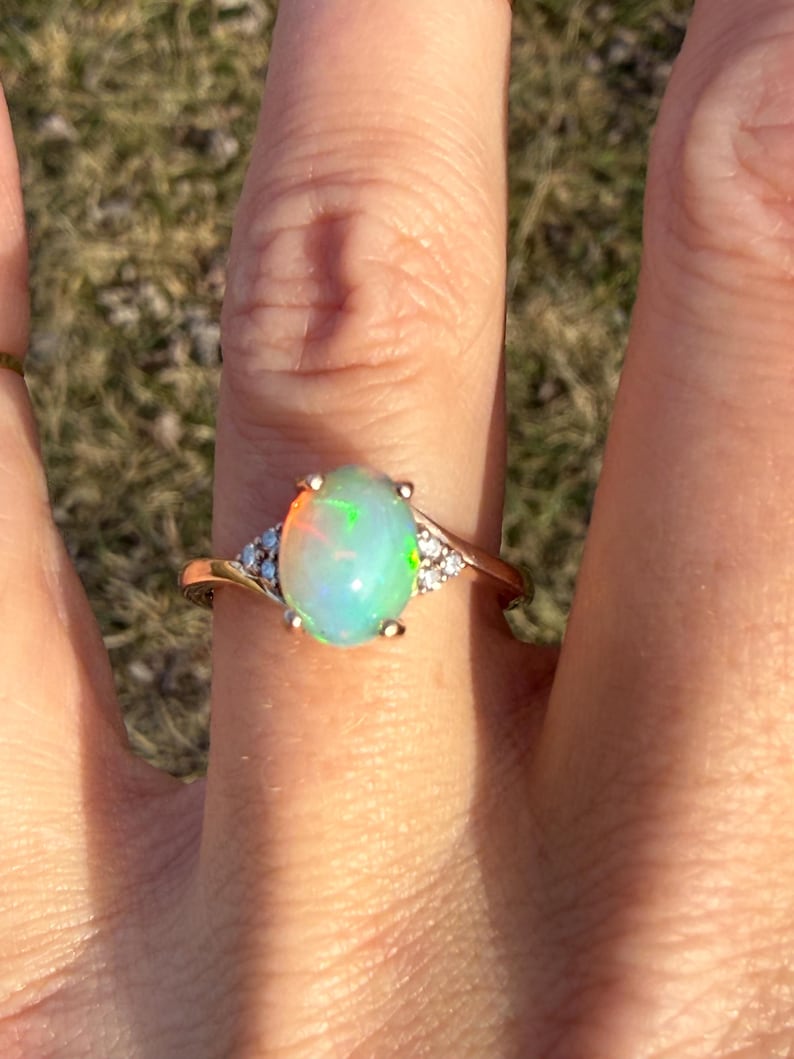 Puede incluir: Un anillo con un cabuj&oacute;n de &oacute;palo ovalado con destellos iridiscentes en tonos verde, naranja y azul. El &oacute;palo est&aacute; engastado en una banda de oro pulido, decorada con tres peque&ntilde;as piedras redondas y transparentes a cada lado.