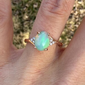 Puede incluir: Un anillo de oro con un &oacute;palo central en forma de cabuj&oacute;n ovalado que muestra destellos iridiscentes verdes y naranjas. El &oacute;palo est&aacute; sujeto por cuatro garras, flanqueado a cada lado por tres peque&ntilde;as piedras redondas claras en el aro.