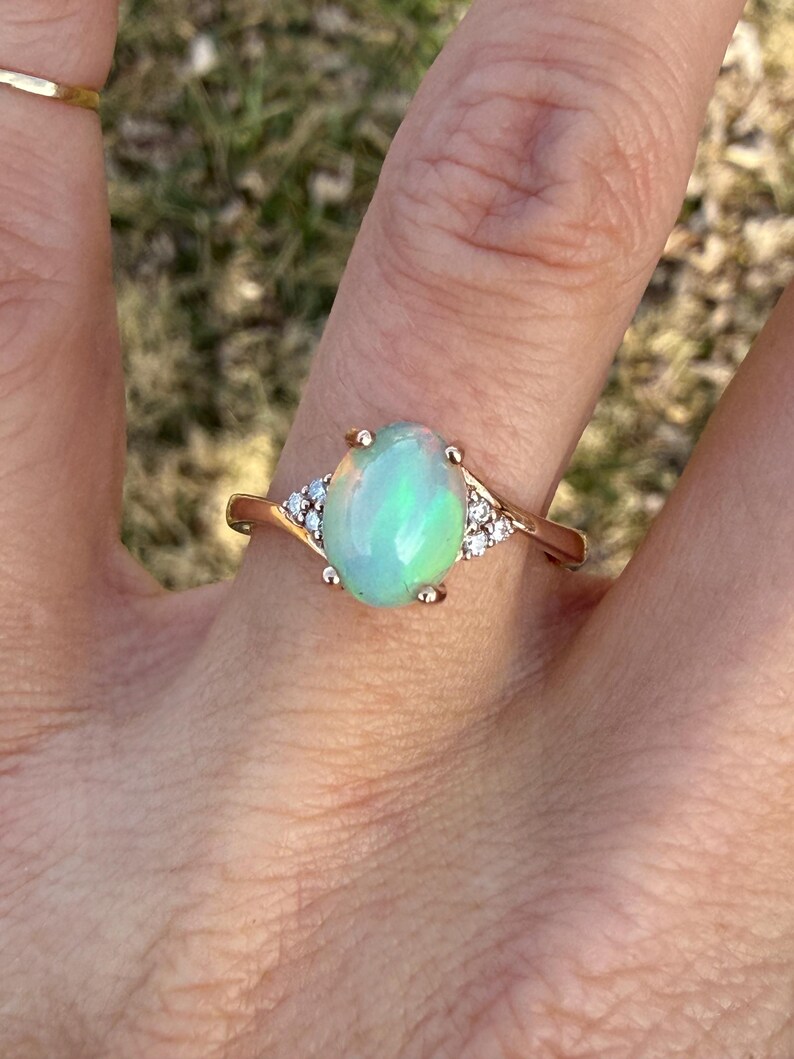 Puede incluir: Un anillo de oro con un &oacute;palo central en forma de cabuj&oacute;n ovalado que muestra destellos iridiscentes verdes y naranjas. El &oacute;palo est&aacute; sujeto por cuatro garras, flanqueado a cada lado por tres peque&ntilde;as piedras redondas claras en el aro.