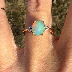 Puede incluir: Un anillo de oro con un &oacute;palo central de forma ovalada con destellos iridiscentes en tonos verde, naranja y azul. El &oacute;palo est&aacute; sujeto por un engaste de cuatro garras, flanqueado por tres peque&ntilde;as piedras redondas transparentes a cada lado.