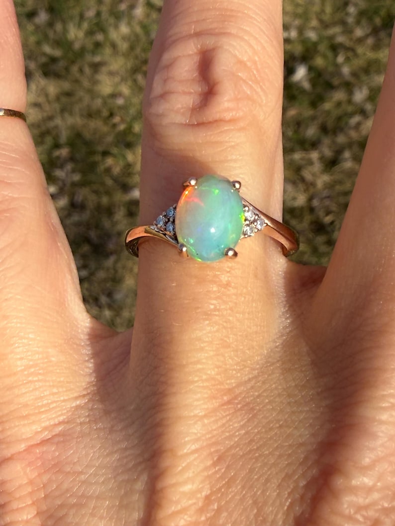 Puede incluir: Un anillo de oro con un &oacute;palo central de forma ovalada con destellos iridiscentes en tonos verde, naranja y azul. El &oacute;palo est&aacute; sujeto por un engaste de cuatro garras, flanqueado por tres peque&ntilde;as piedras redondas transparentes a cada lado.