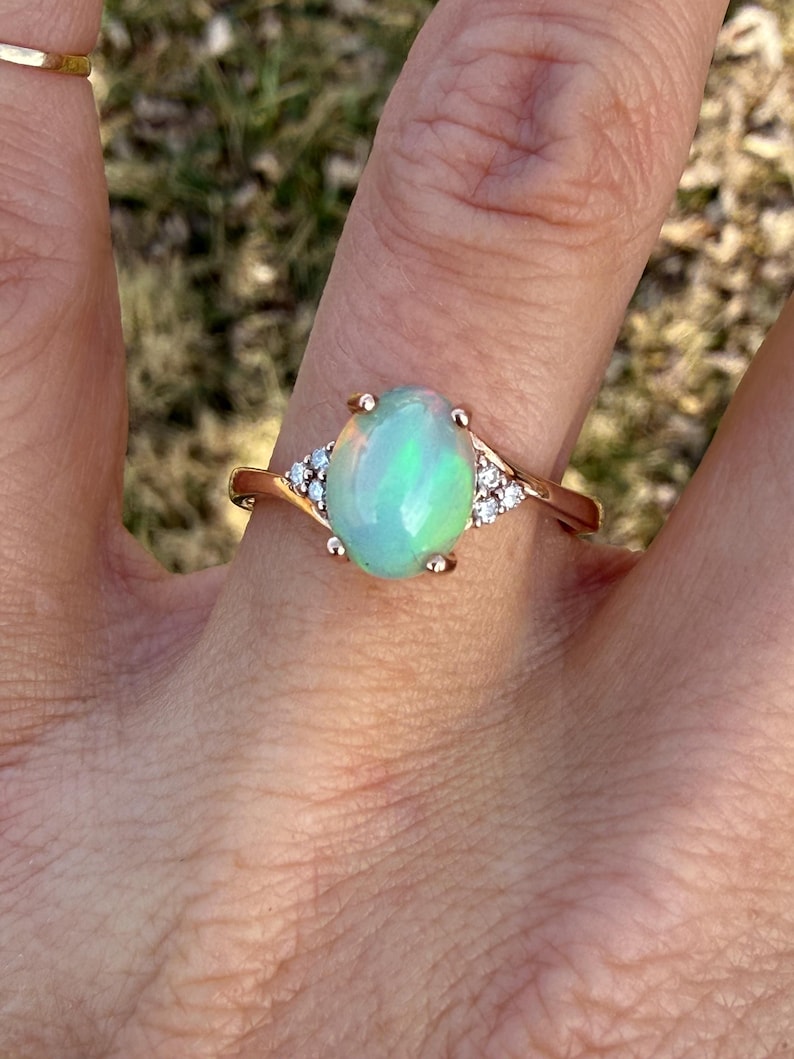 Puede incluir: Un anillo con un &oacute;palo cabuj&oacute;n ovalado montado en una banda de oro rosa pulido. El &oacute;palo central muestra destellos iridiscentes de color verde y naranja. Tres peque&ntilde;as piedras redondas y transparentes est&aacute;n engastadas a cada lado.