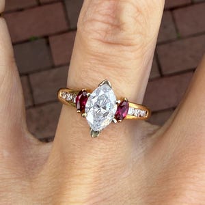 Può includere: Un anello d'oro con un grande diamante centrale a taglio marquise, affiancato da due pietre rosse ovali più piccole. La fascia è inoltre impreziosita da diamanti più piccoli. L'anello è mostrato su un dito.