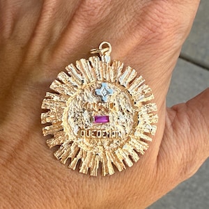 May include: A round gold pendant with a textured sunburst border. The center features the French text 'QU'HIER QUE DEMAIN' in raised letters, a small silver cross with a central diamond, and a rectangular pink gemstone set horizontally.