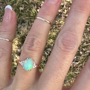 Puede incluir: Un anillo de oro con una piedra central de &oacute;palo ovalada con un sutil juego de colores iridiscentes, flanqueada por tres peque&ntilde;as piedras transparentes a cada lado. La banda es fina y de oro pulido, usada junto a dos anillos apilables simples.