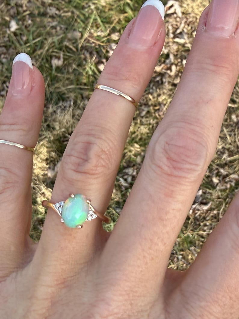 Puede incluir: Un anillo de oro con una piedra central de &oacute;palo ovalada con un sutil juego de colores iridiscentes, flanqueada por tres peque&ntilde;as piedras transparentes a cada lado. La banda es fina y de oro pulido, usada junto a dos anillos apilables simples.