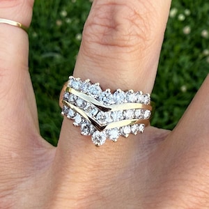 May include: A gold ring with multiple rows of clear, sparkling gemstones. The ring has a layered design with a chevron-shaped top. The gemstones are set in a gold band, creating a luxurious and eye-catching piece of jewelry.