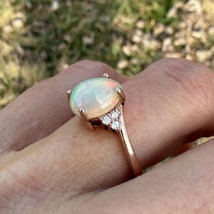 Puede incluir: Un anillo de oro rosa con un gran &oacute;palo cabuj&oacute;n de forma ovalada con destellos iridiscentes verdes y naranjas. El &oacute;palo est&aacute; sujeto por cuatro garras y la banda est&aacute; adornada con tres peque&ntilde;as piedras redondas y transparentes a cada lado.