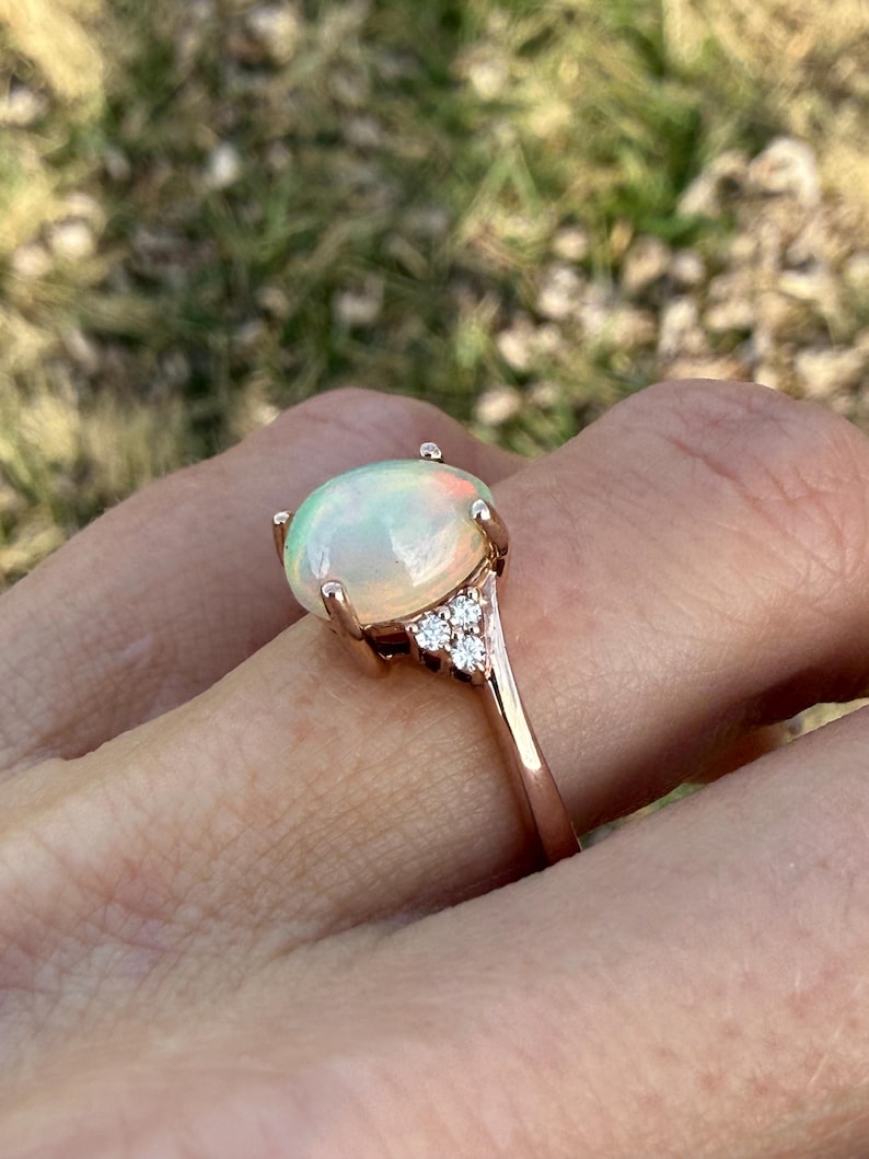 Puede incluir: Un anillo de oro rosa con un gran &oacute;palo cabuj&oacute;n de forma ovalada con destellos iridiscentes verdes y naranjas. El &oacute;palo est&aacute; sujeto por cuatro garras y la banda est&aacute; adornada con tres peque&ntilde;as piedras redondas y transparentes a cada lado.