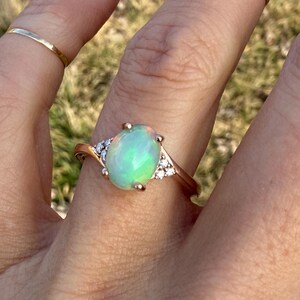 Puede incluir: Un anillo de oro rosa con un &oacute;palo central de forma ovalada con destellos iridiscentes verdes y naranjas, flanqueado por tres peque&ntilde;as piedras redondas transparentes a cada lado. El aro es liso y pulido, acompa&ntilde;ado de dos anillos finos.