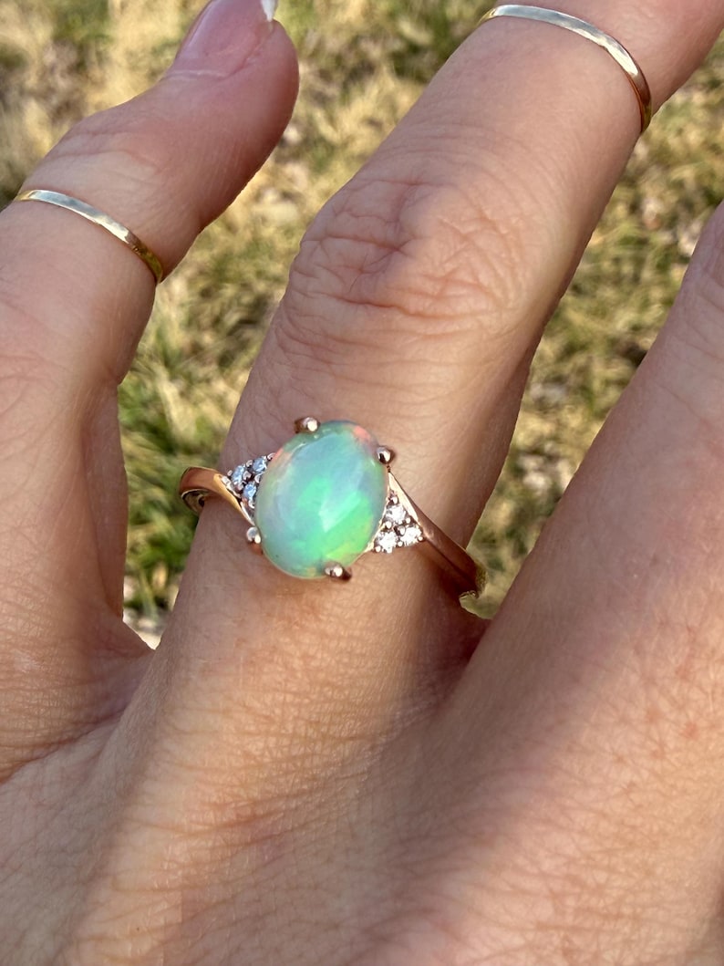 Puede incluir: Un anillo de oro rosa con un &oacute;palo central de forma ovalada con destellos iridiscentes verdes y naranjas, flanqueado por tres peque&ntilde;as piedras redondas transparentes a cada lado. El aro es liso y pulido, acompa&ntilde;ado de dos anillos finos.