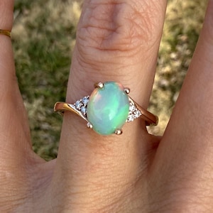 Puede incluir: Un anillo con un &oacute;palo cabuj&oacute;n de forma ovalada engastado en una banda de metal pulido. El &oacute;palo central muestra destellos iridiscentes verdes y naranjas. El anillo presenta un engaste estilo bypass con tres peque&ntilde;as piedras redondas transparentes a cada lado.