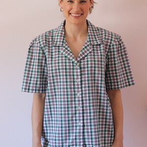 May include: A woman wearing a white, green, and black plaid short-sleeved button-up shirt with a collared neckline. She is also wearing light blue jeans.