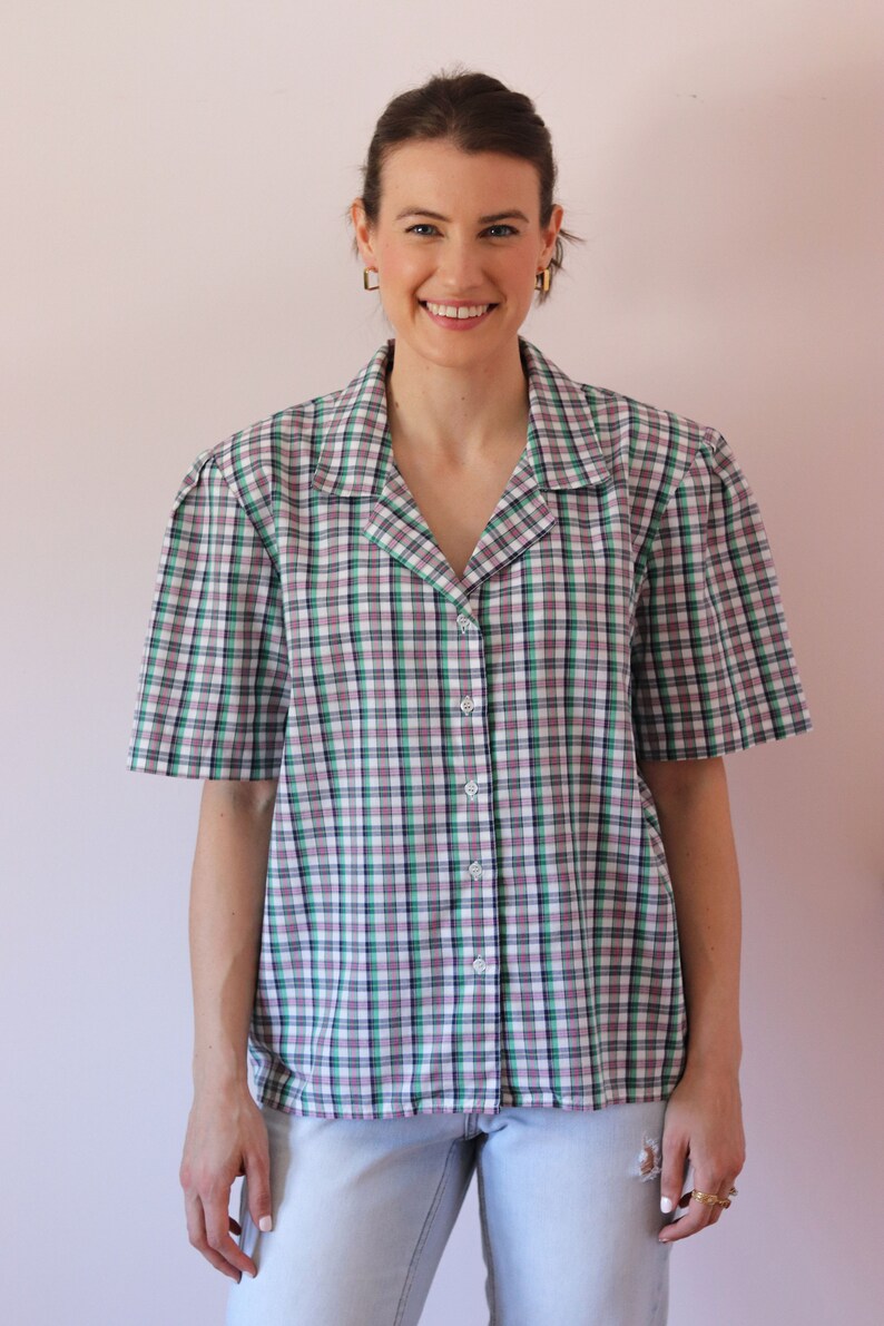 May include: A woman wearing a white, green, and black plaid short-sleeved button-up shirt with a collared neckline. She is also wearing light blue jeans.