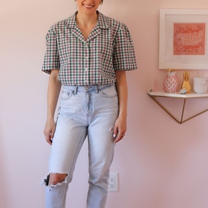 May include: A woman wearing a green and white plaid short-sleeved shirt with a light blue pair of jeans and white sneakers. The shirt has a collar and buttons down the front. The jeans have a distressed look with a hole in one knee. The woman is standing in front of a pink wall with a framed print that says "Nothing Changes Until Nothing Changes".