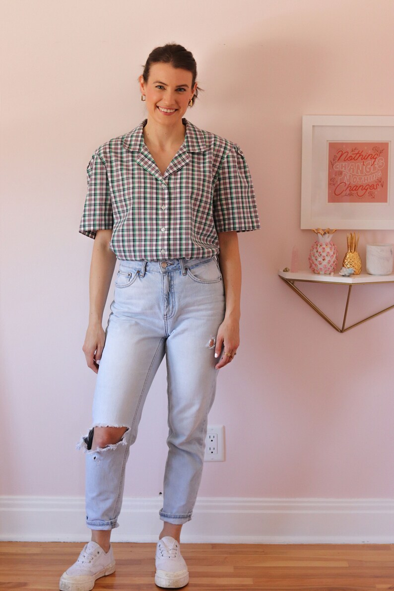 May include: A woman wearing a green and white plaid short-sleeved shirt with a light blue pair of jeans and white sneakers. The shirt has a collar and buttons down the front. The jeans have a distressed look with a hole in one knee. The woman is standing in front of a pink wall with a framed print that says "Nothing Changes Until Nothing Changes".